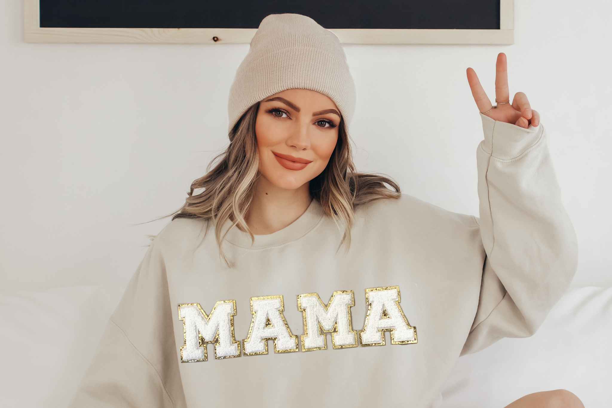 Woman wearing a beige sweatshirt with 'MAMA' chenille letters, making a peace sign.