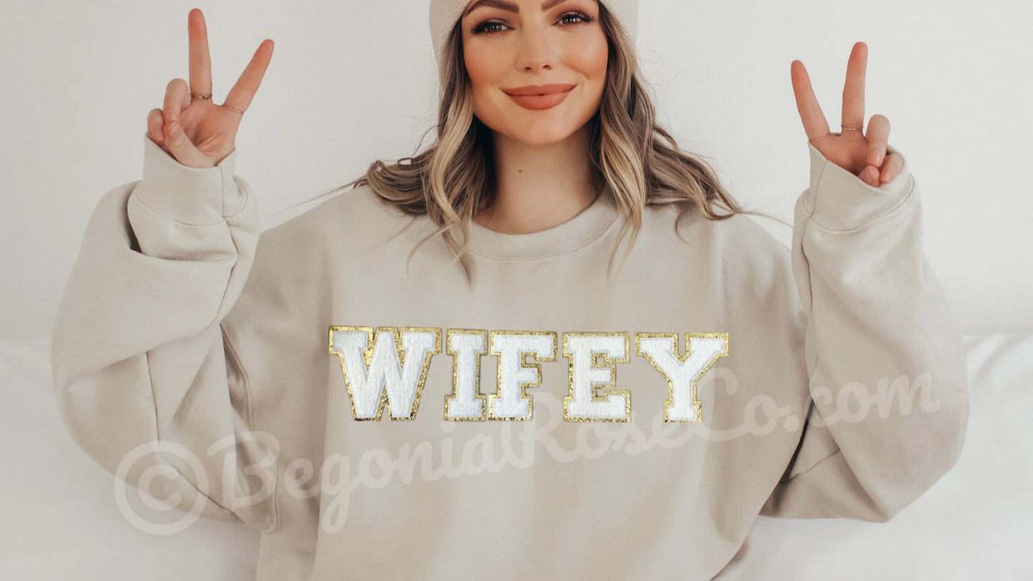 Woman wearing a beige sweatshirt with 'WIFEY' in gold letters, making peace signs.
