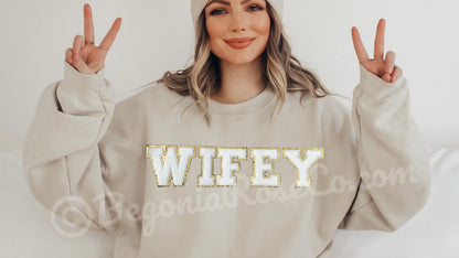 Woman wearing a beige sweatshirt with 'WIFEY' in gold letters, making peace signs.
